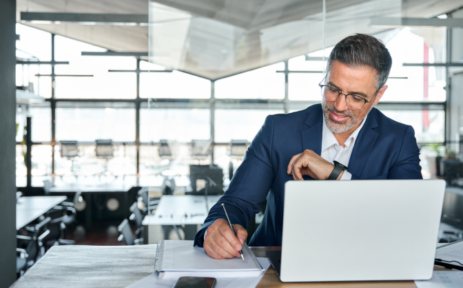 Foto de un hombre adulto con gafas y traje escribiendo en un documento y consultando en su computador portátil cómo es la transparencia y acceso a la información en Valores Bancolombia.