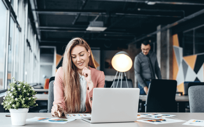 Foto de una mujer joven con cabello largo, sonriendo en su escritorio donde está su computador portátil y mirando los documentos con los resultados de la inversión inmobiliaria con Valores Bancolombia.