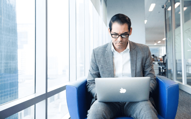 Foto de un hombre con gafas y traje sentado en un mueble con un computador portátil en sus piernas leyendo sobre las alternativas de inversión en Valores Bancolombia.